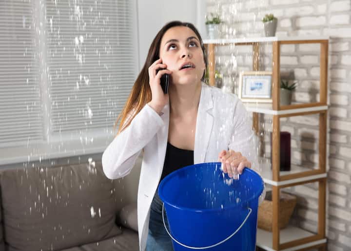 Water-Damage-Leak A woman calling water damage repair experts while holding a bucket trying to catch the water leaking from her ceiling in King of Prussia